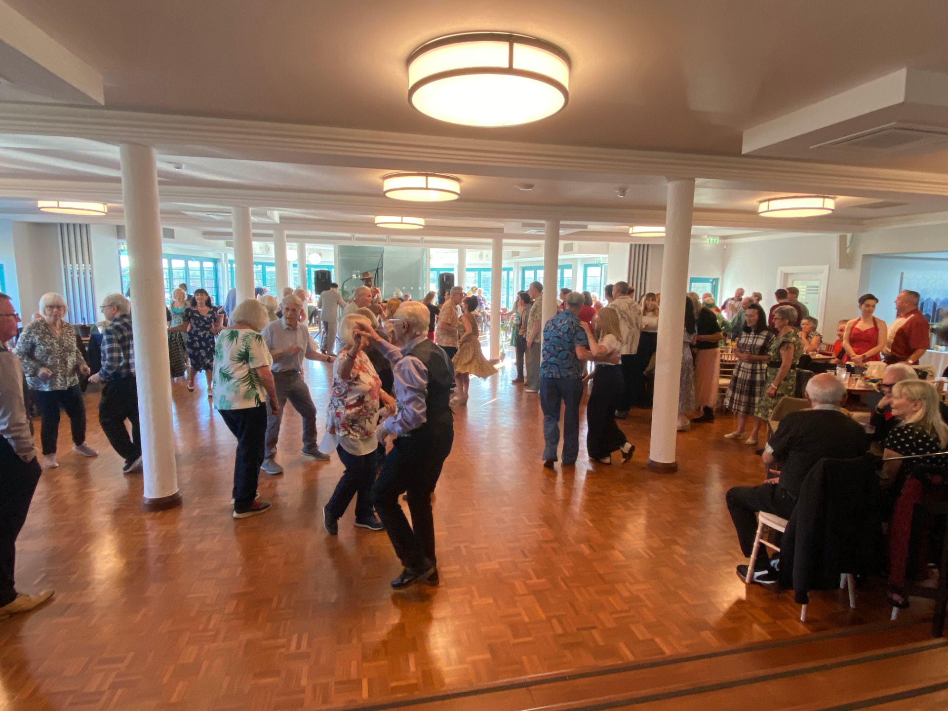 A busy parquet dance floor with couples swing dancing in a bright 1930s seaside ballroom